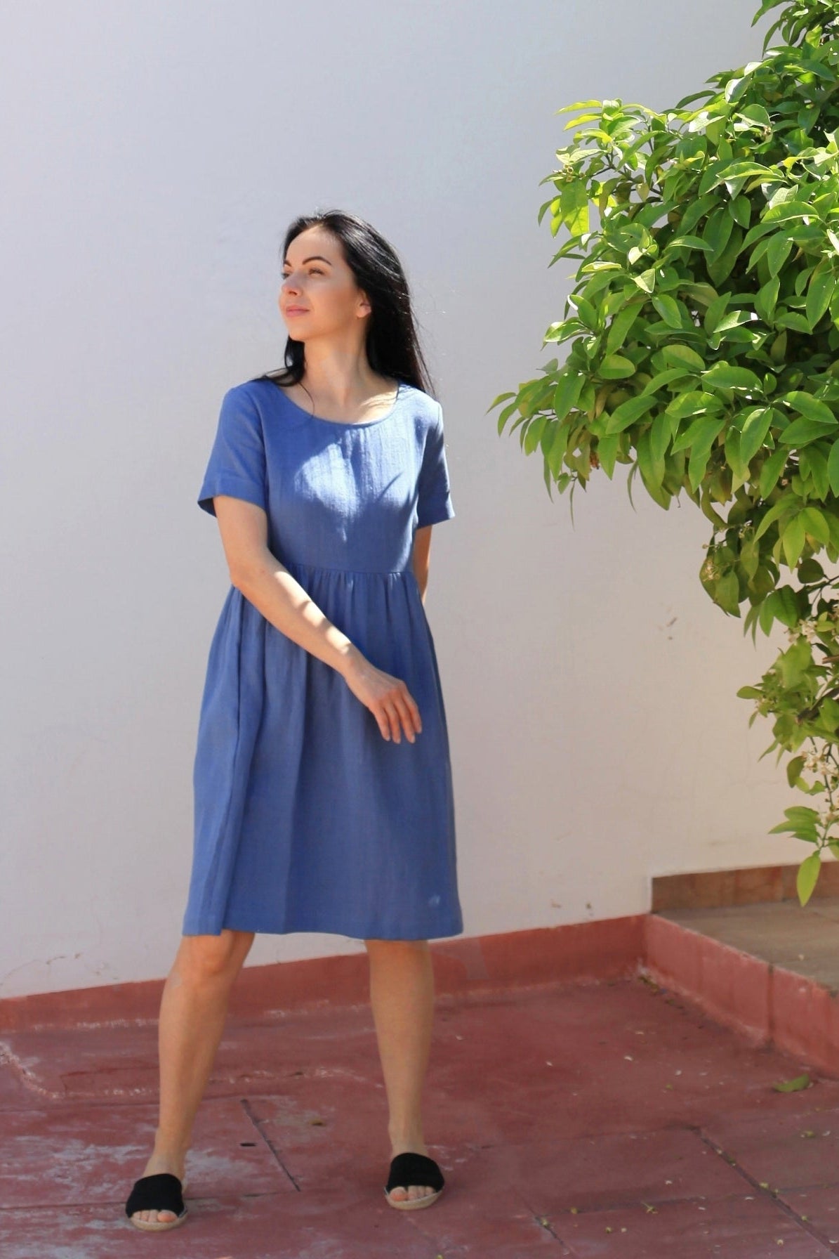 Woman in a blue dress standing next to a tree on a sidewalk.