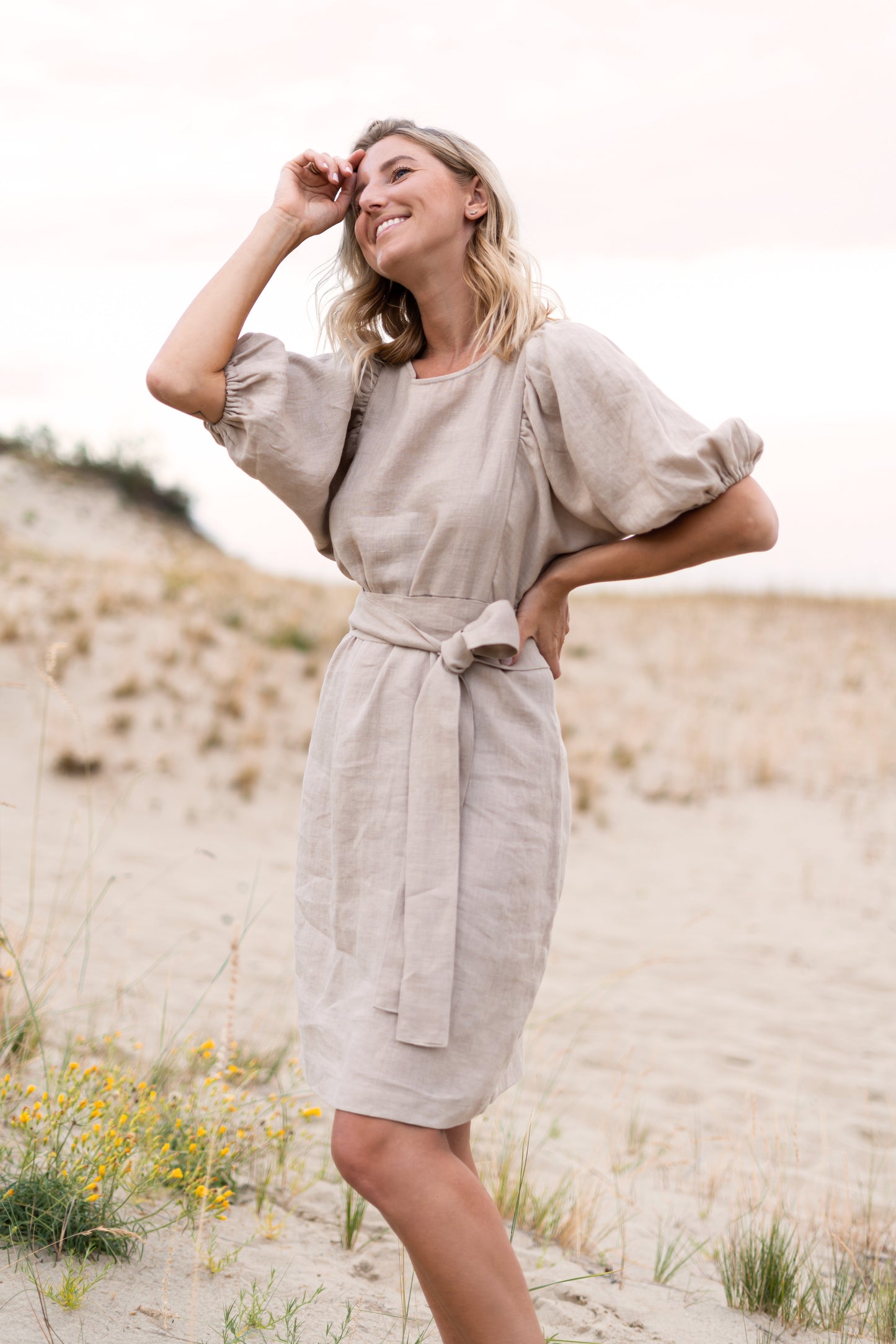 Woman in a beige dress standing on sandy dunes with wildflowers.