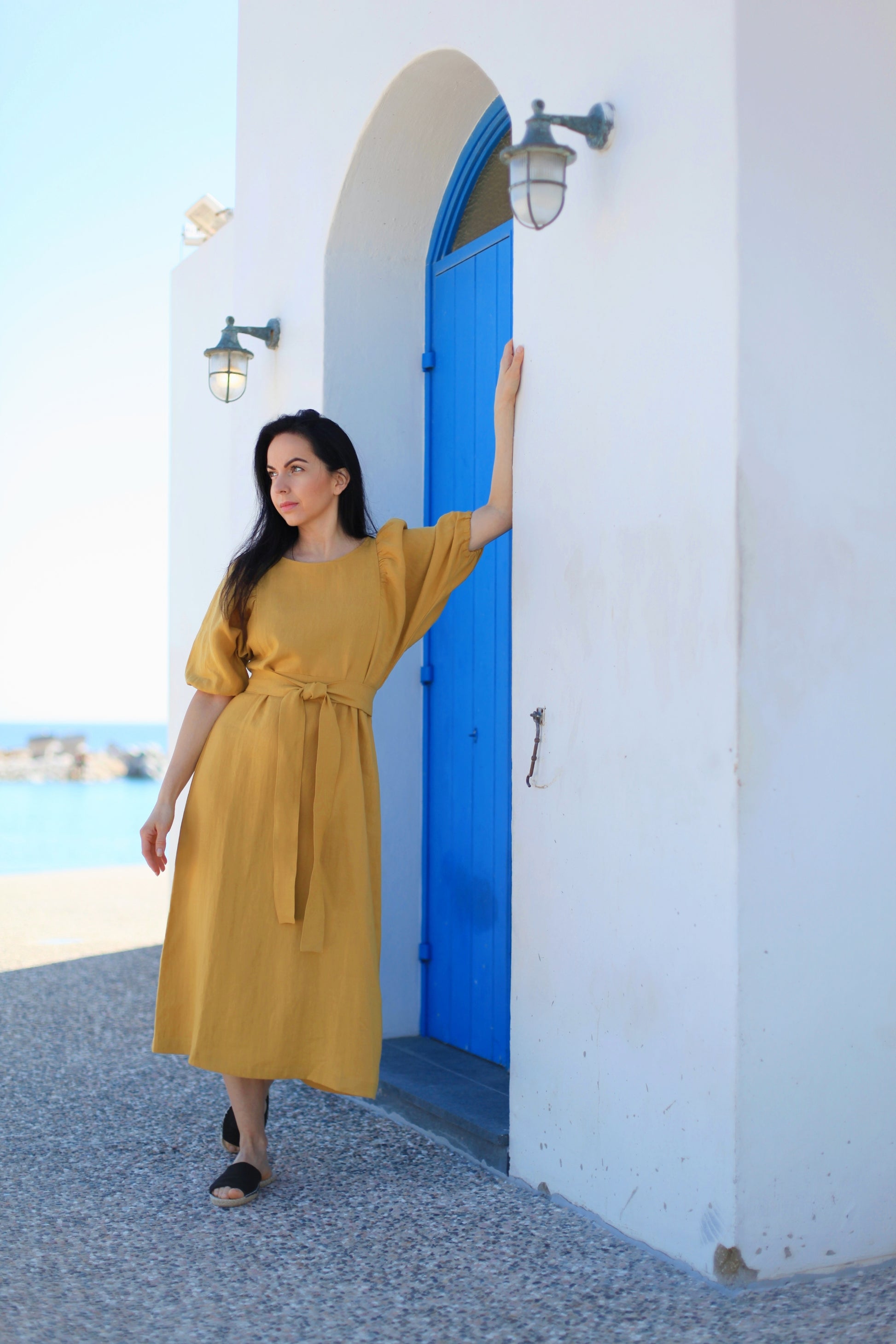 Woman in a yellow dress standing in front of a blue door and white wall with ocean view.