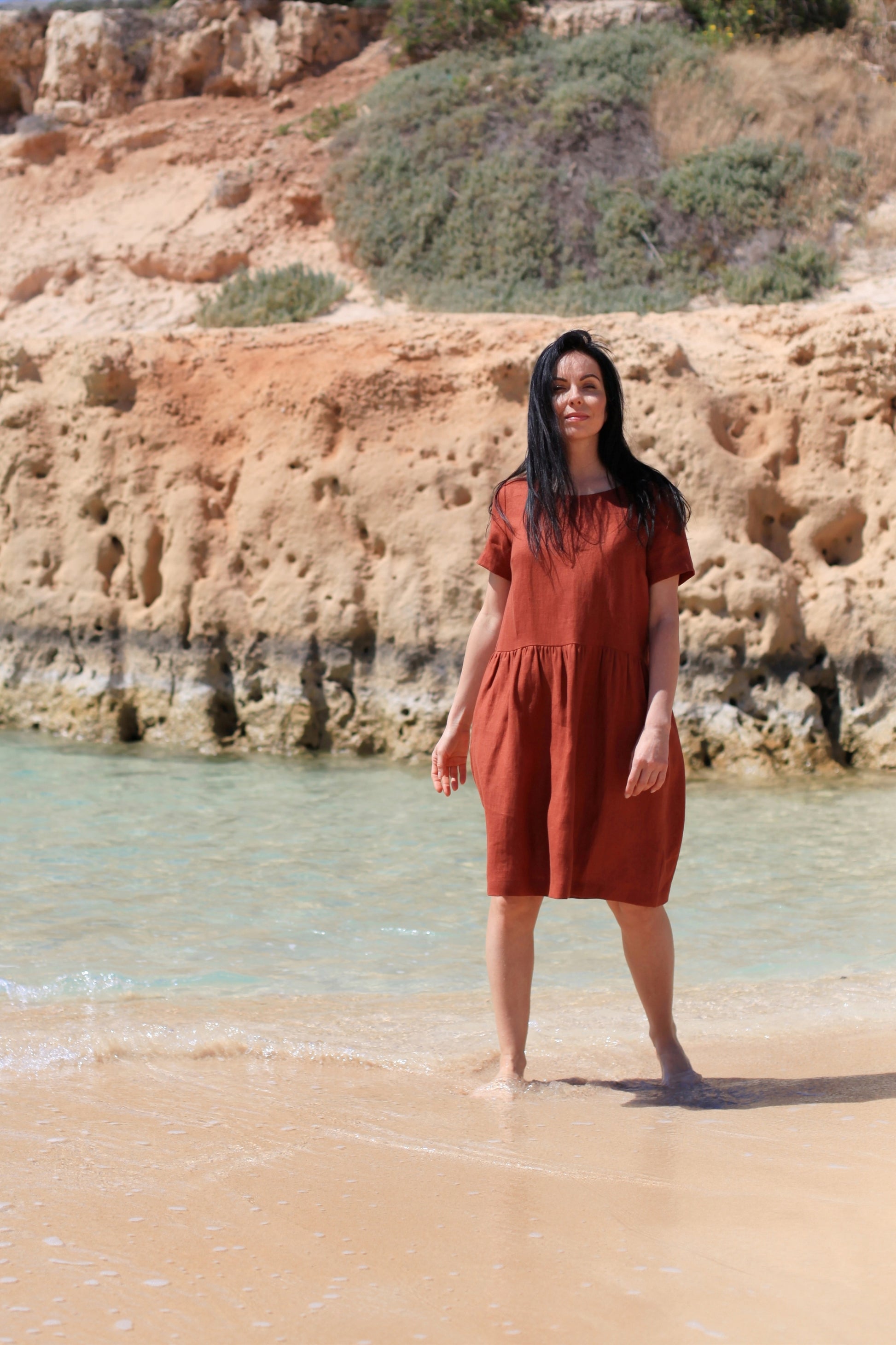 Woman in a rust-colored dress standing on a beach with rocky cliffs in the background