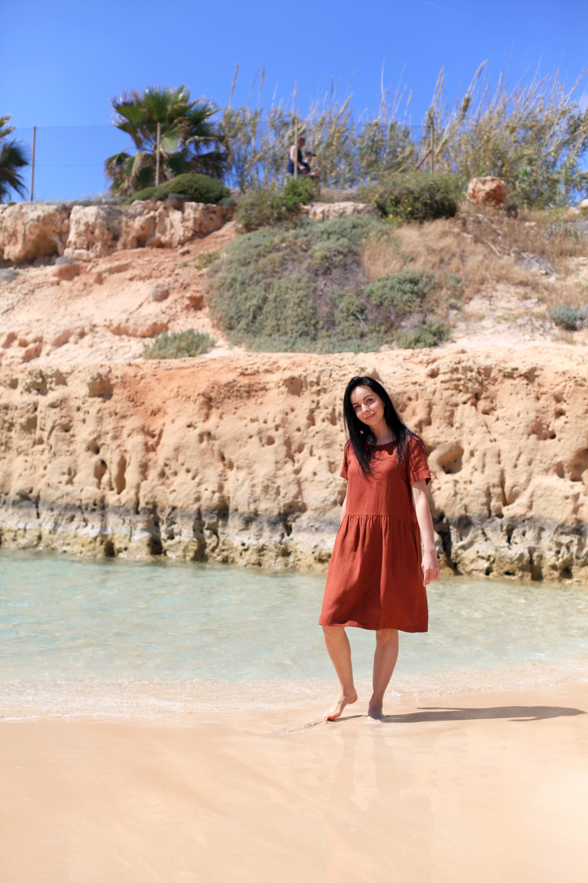 Woman in a red dress standing on a beach with a rocky cliff and palm trees in the background.