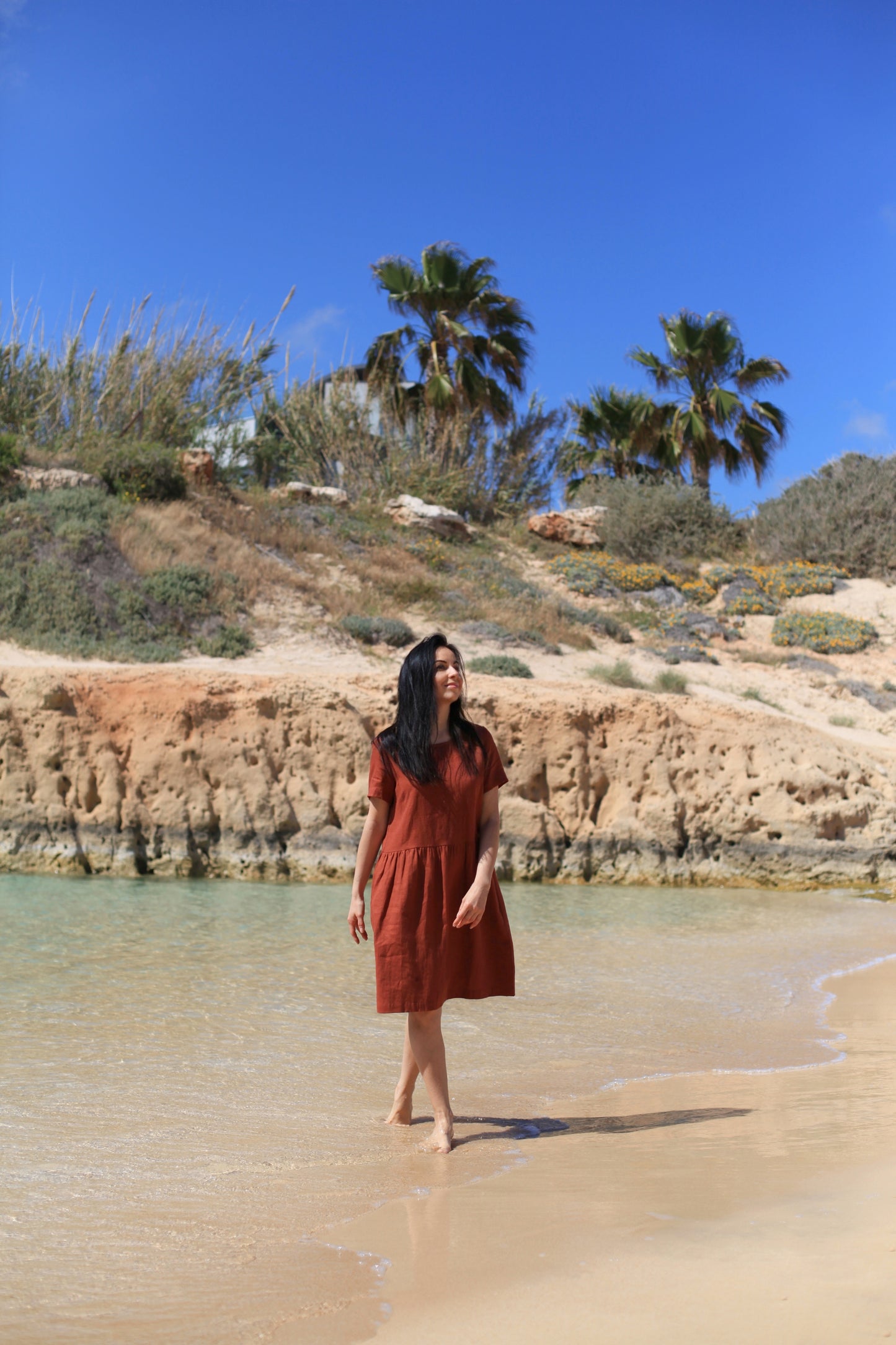 Woman in a red dress standing on a sandy beach with a rocky hill and palm trees in the background.