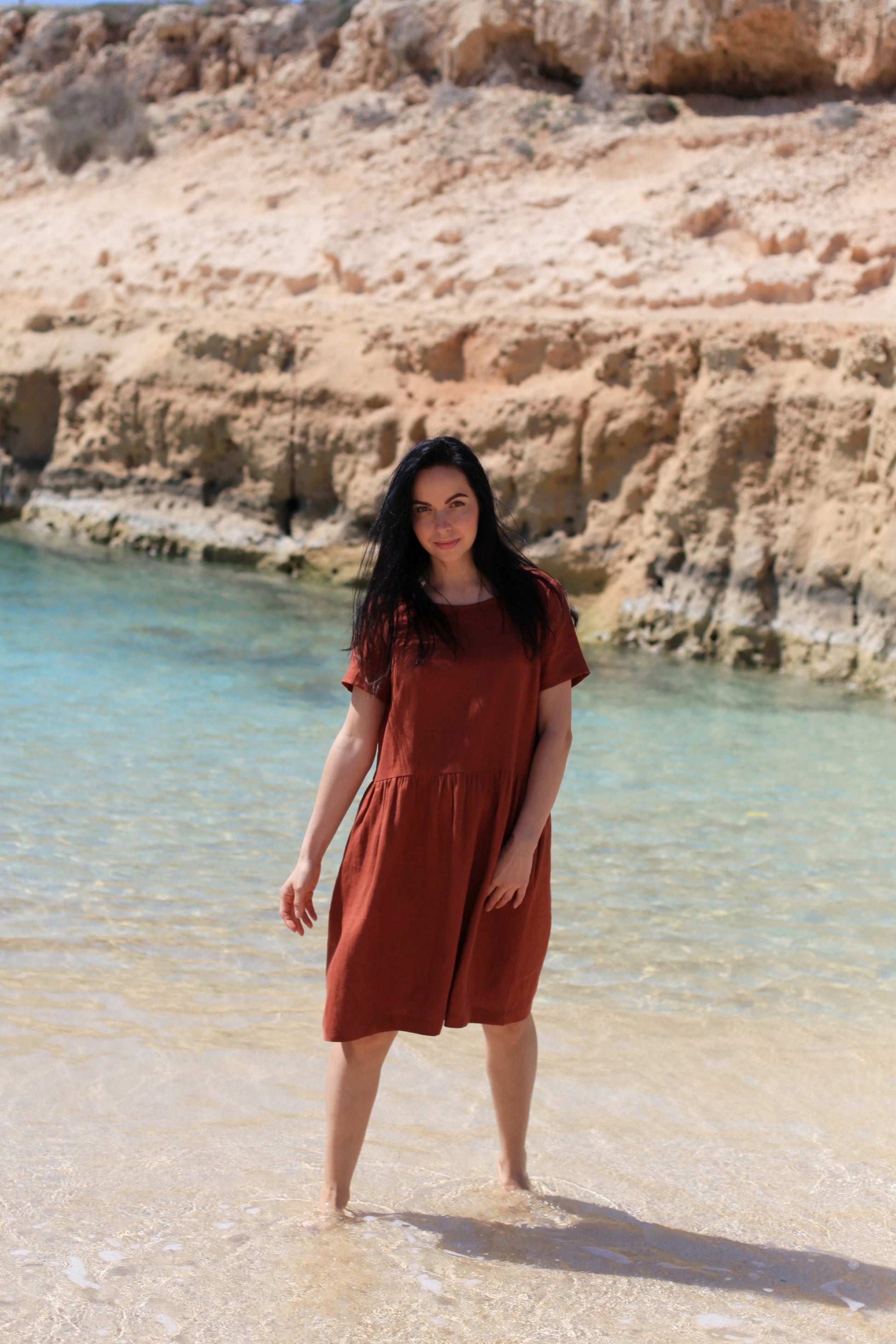 Woman in a rust-colored dress standing in shallow water near a rocky coastline.