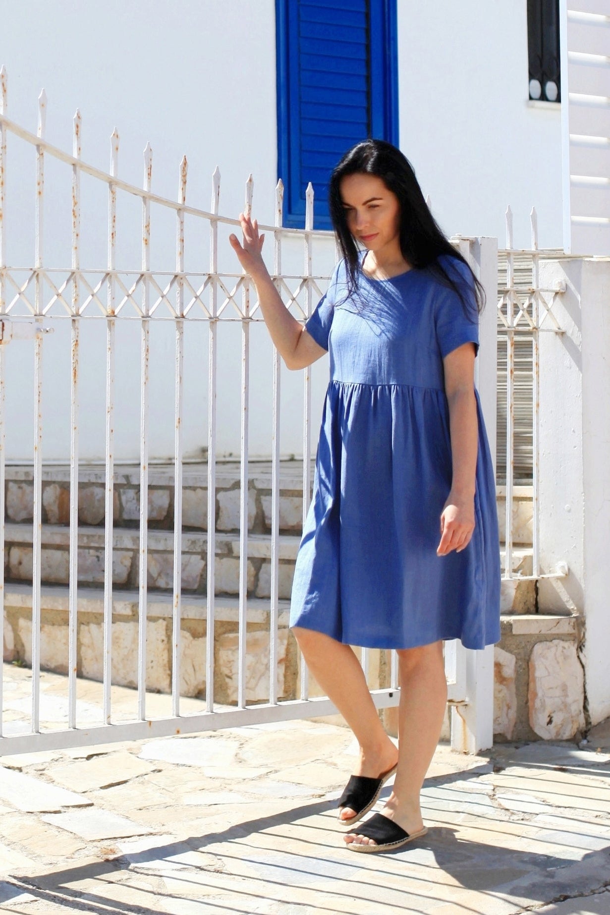 Woman in a blue dress standing on a stone path with a white fence and blue door in the background.