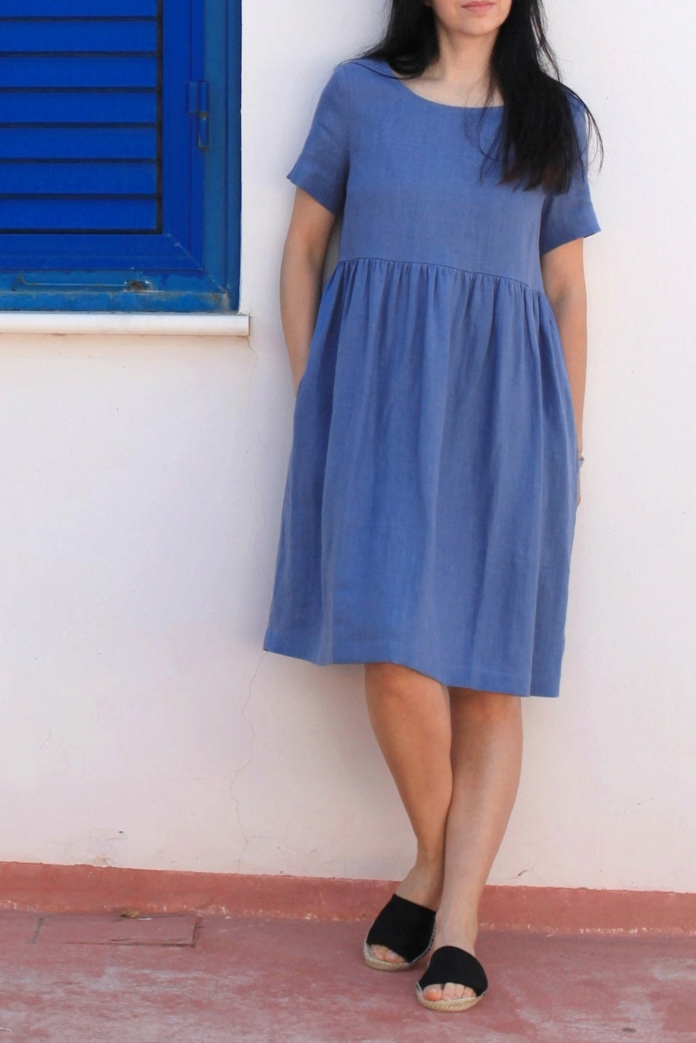 Woman wearing a blue dress standing in front of a white wall with blue shutters.