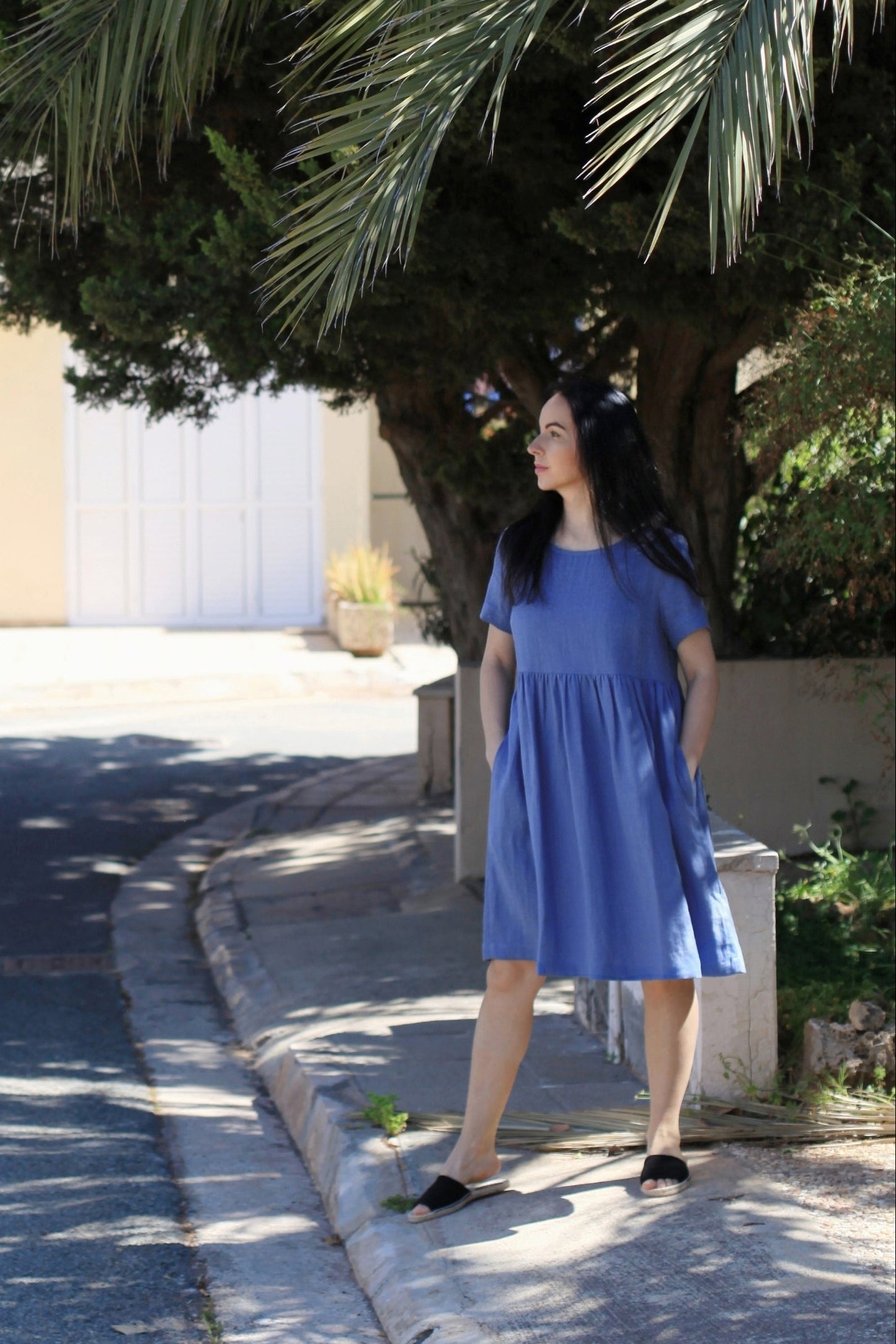 Woman in a blue dress standing on a sidewalk under a tree with a building in the background.