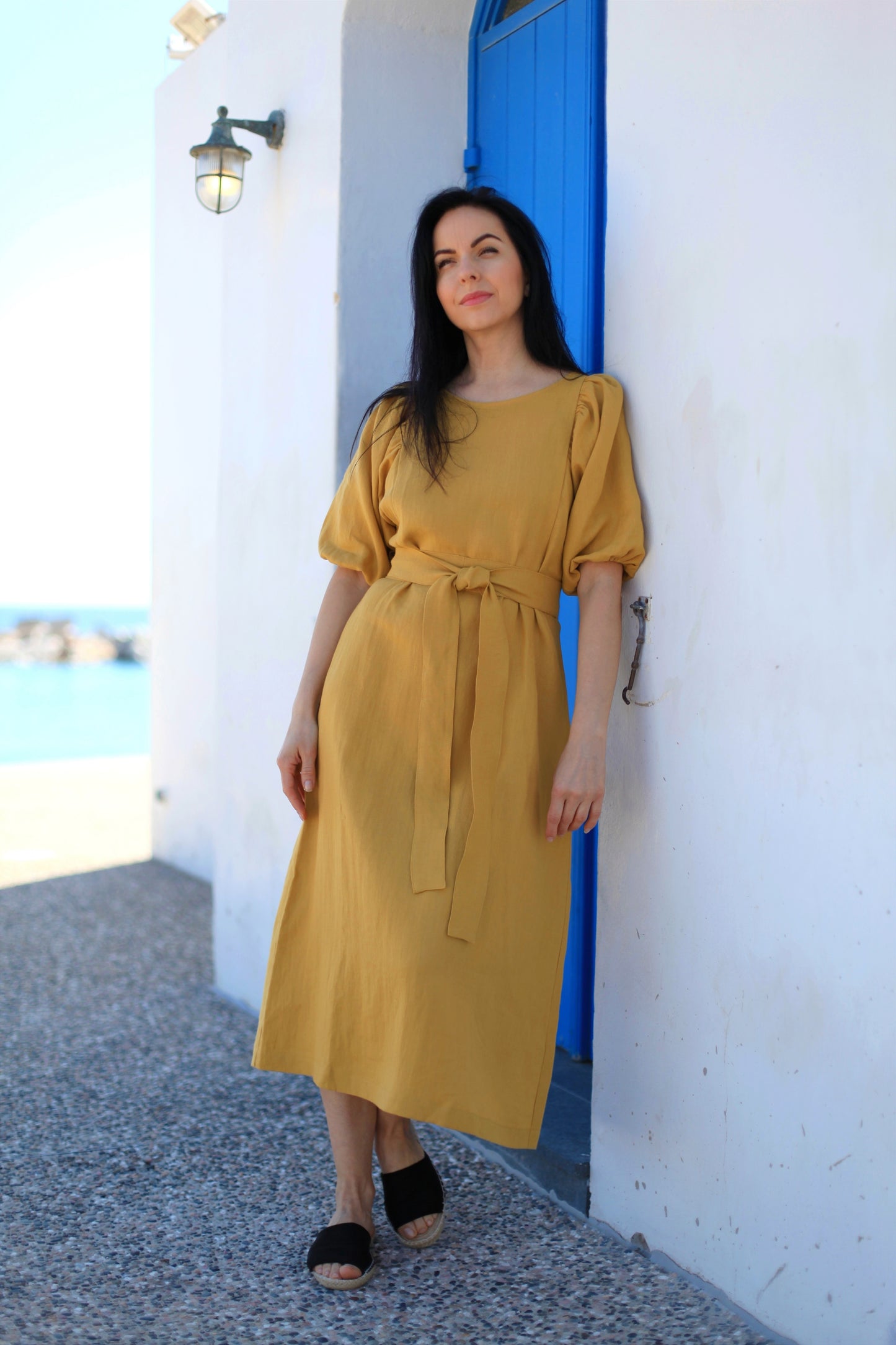 Woman in a yellow dress standing in front of a blue door and white wall with ocean view.