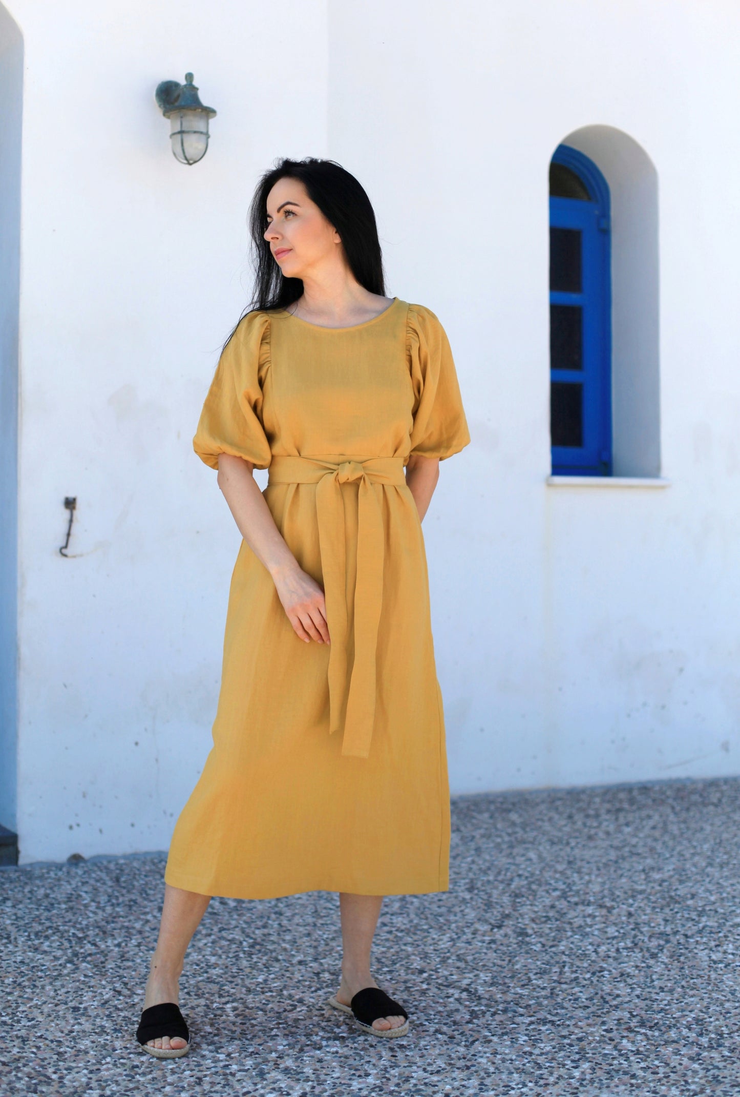Woman wearing a mustard yellow dress standing in front of a white wall with a blue window.