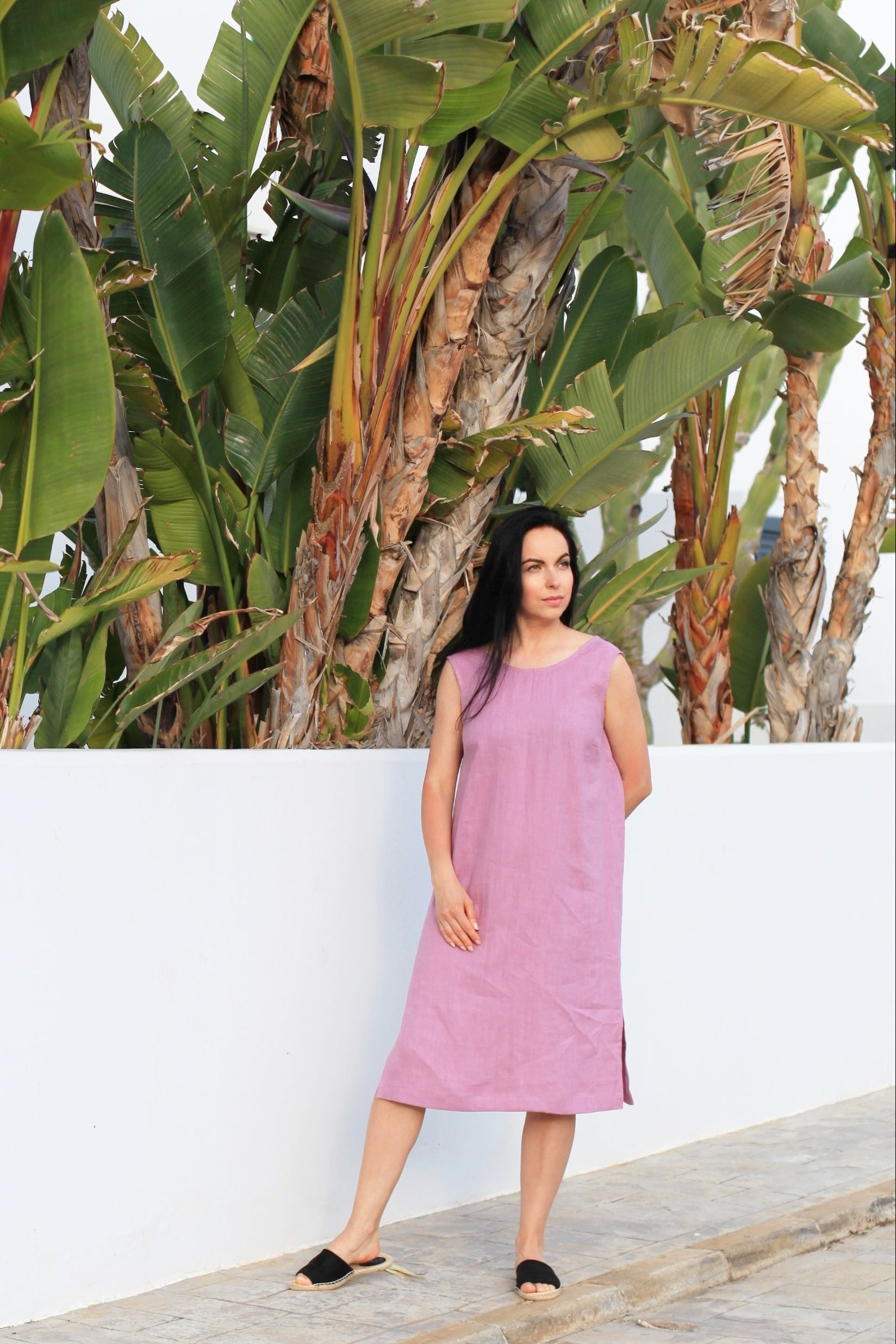Woman in a pink dress standing against a white wall with green plants in the background