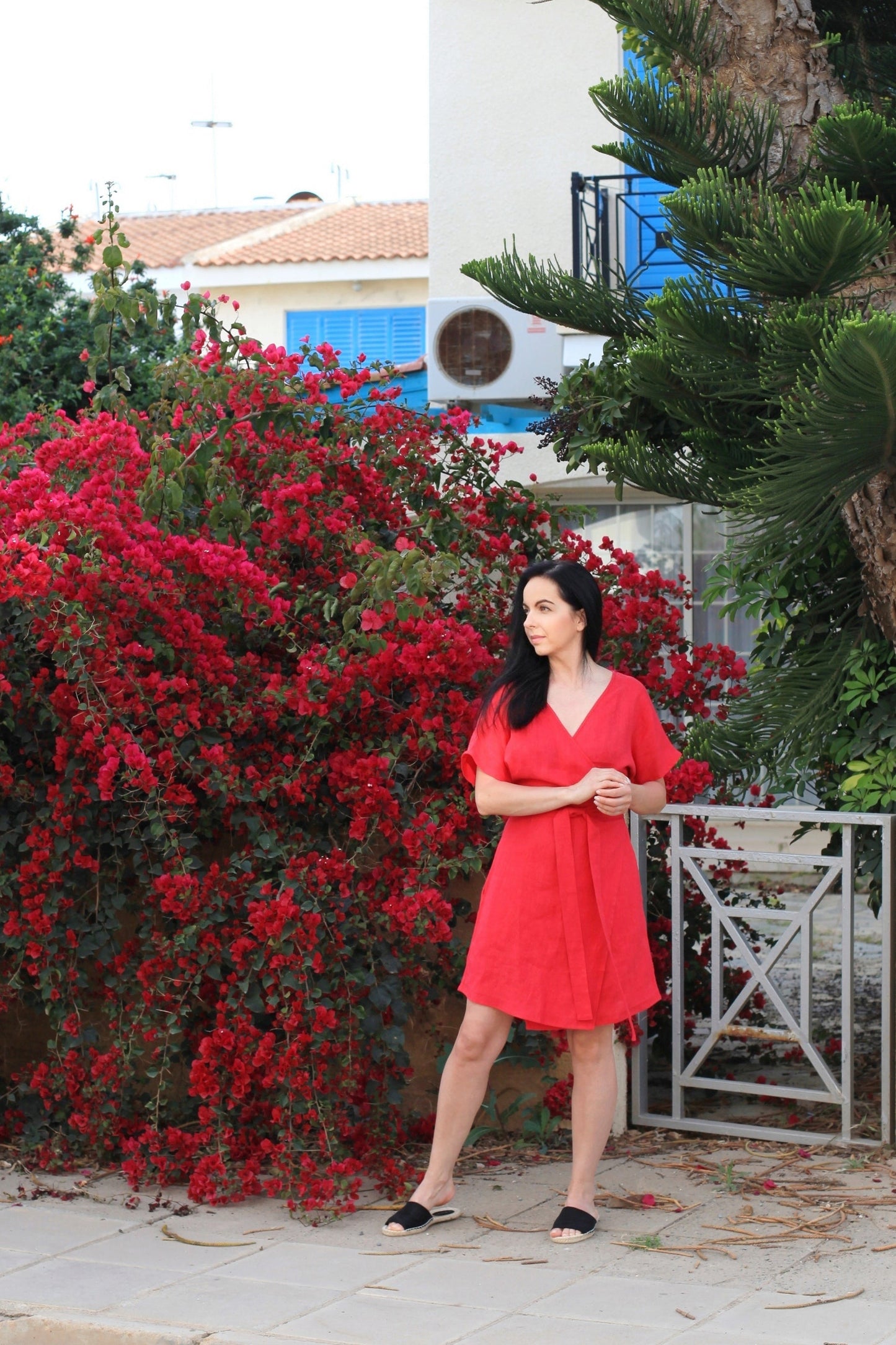 Woman in a red dress standing in front of a bush with red flowers and a building in the background.