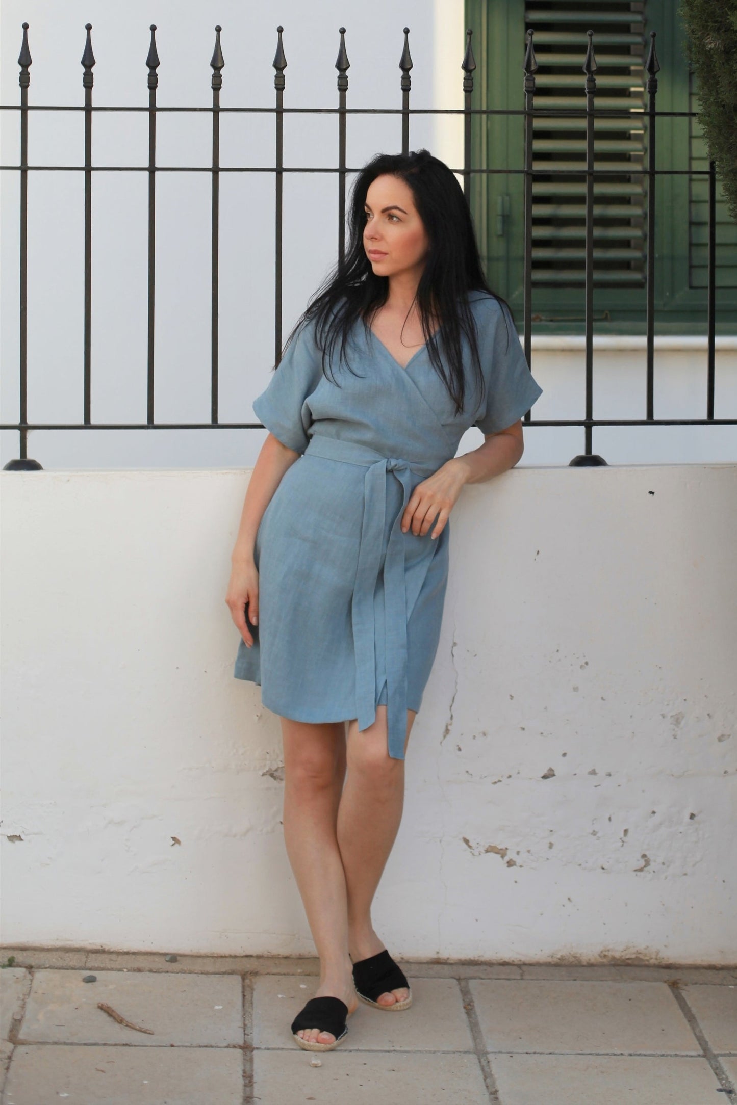 Woman in a blue dress standing against a white wall with a black metal fence and greenery.