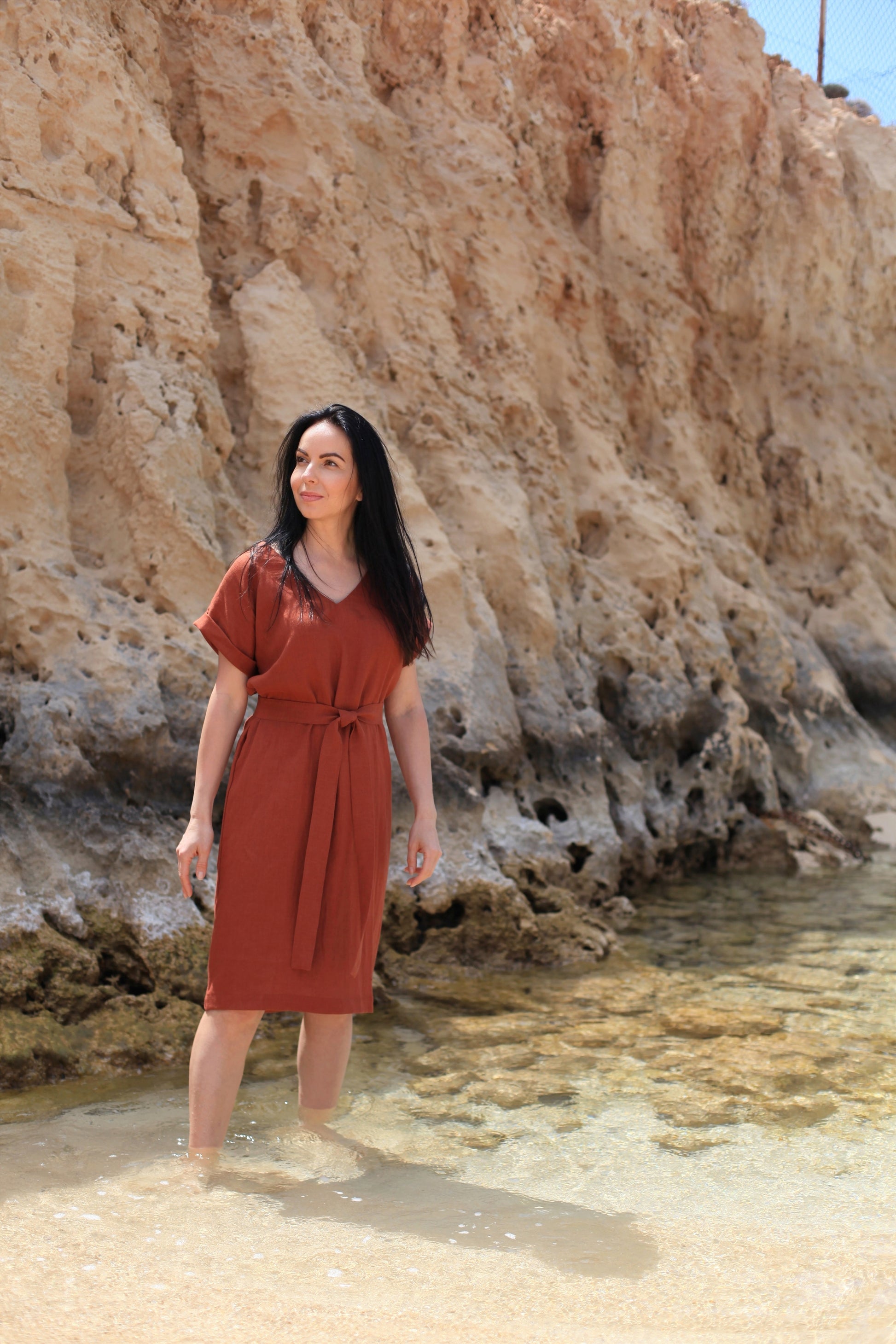 Woman in a rust-colored dress standing in shallow water near rocky cliffs.