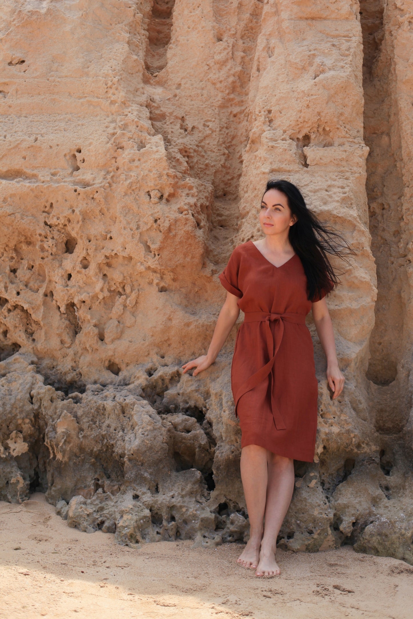 Woman in a rust-colored dress standing against a rocky wall.
