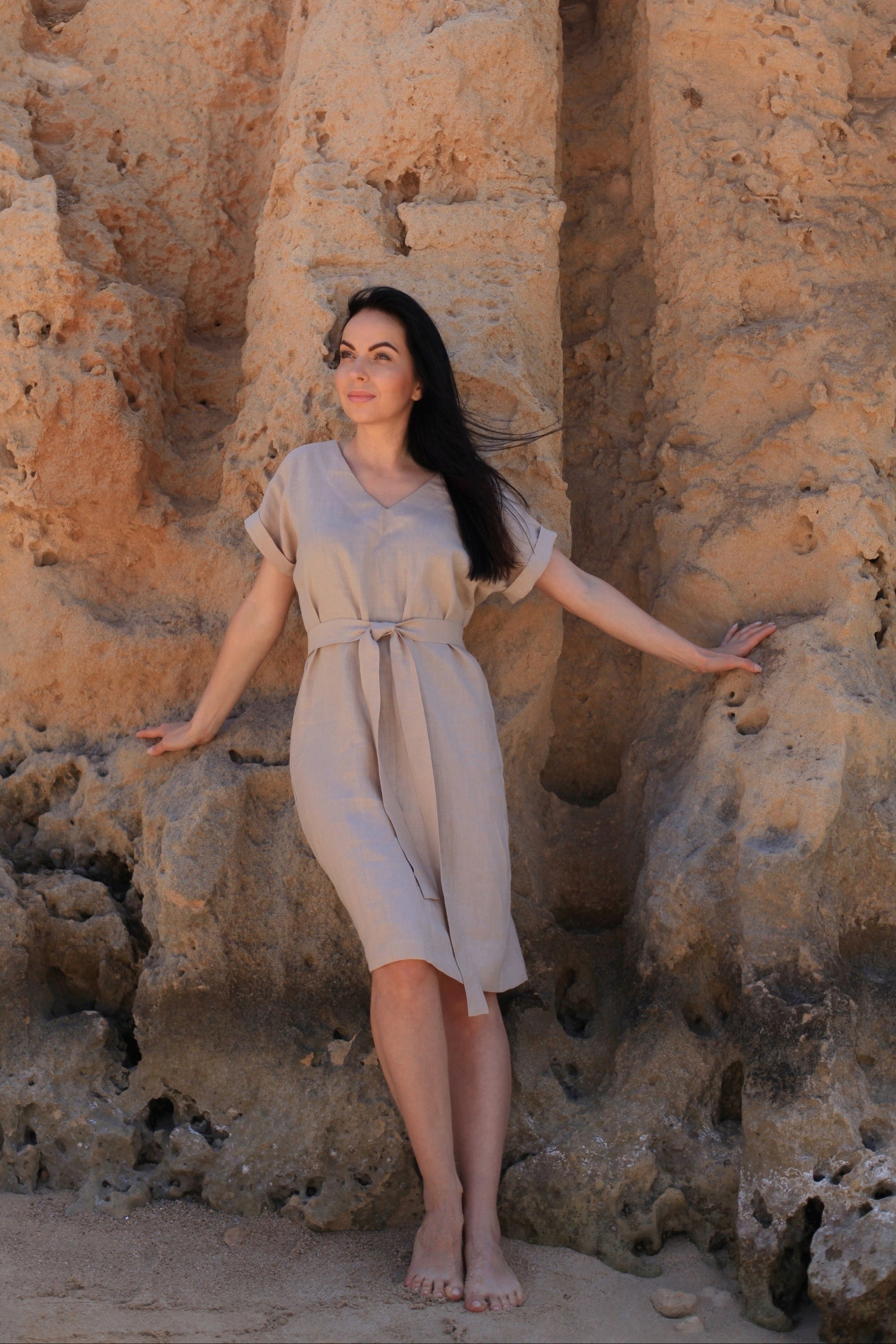 Woman in a beige dress standing against a textured stone wall.