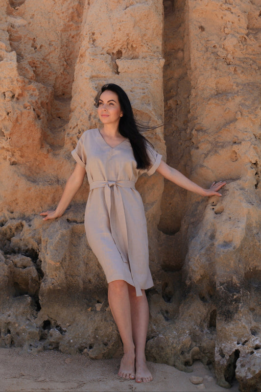 Woman in a beige dress standing against a textured stone wall.