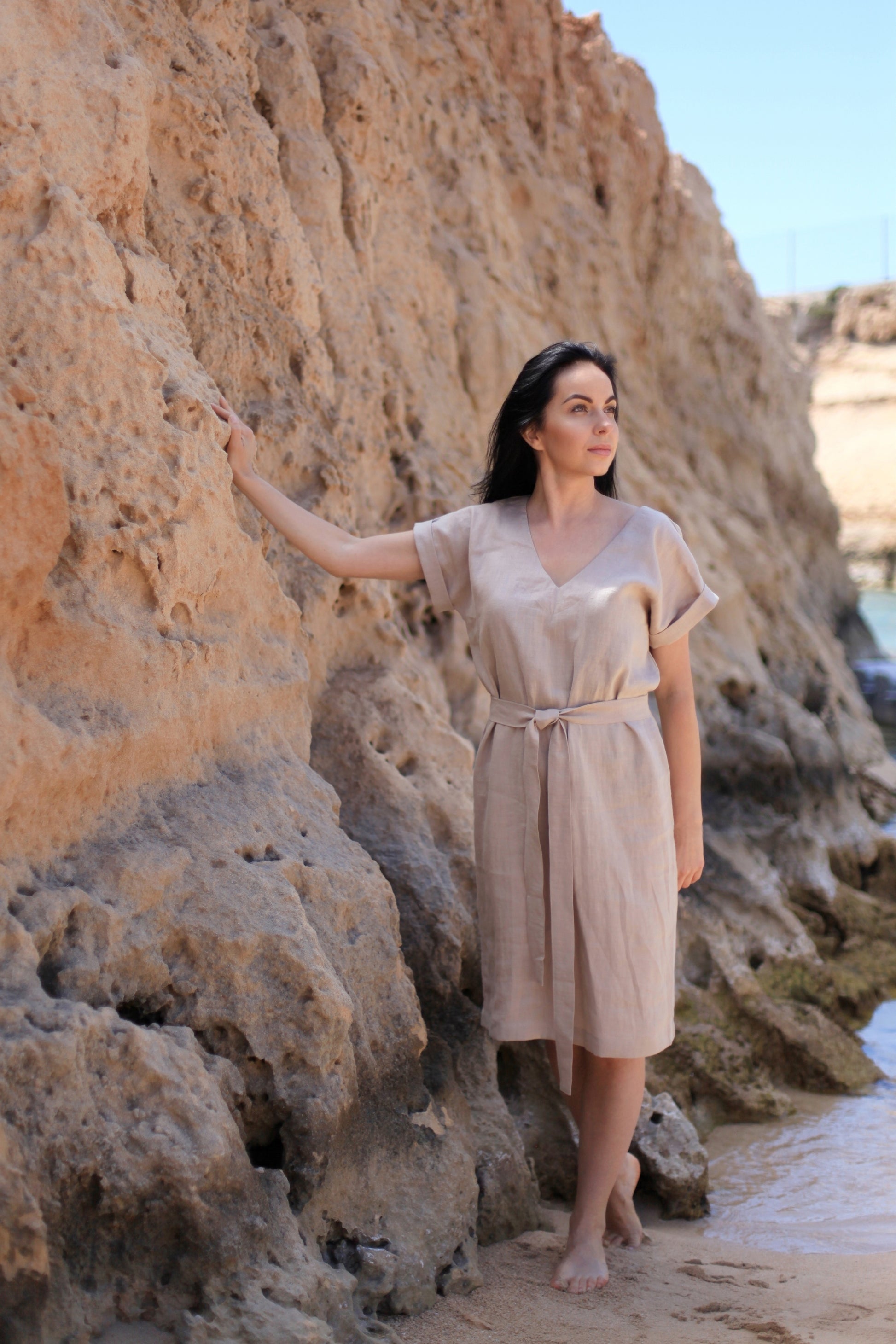 Woman in a beige dress standing against a rocky beach backdrop