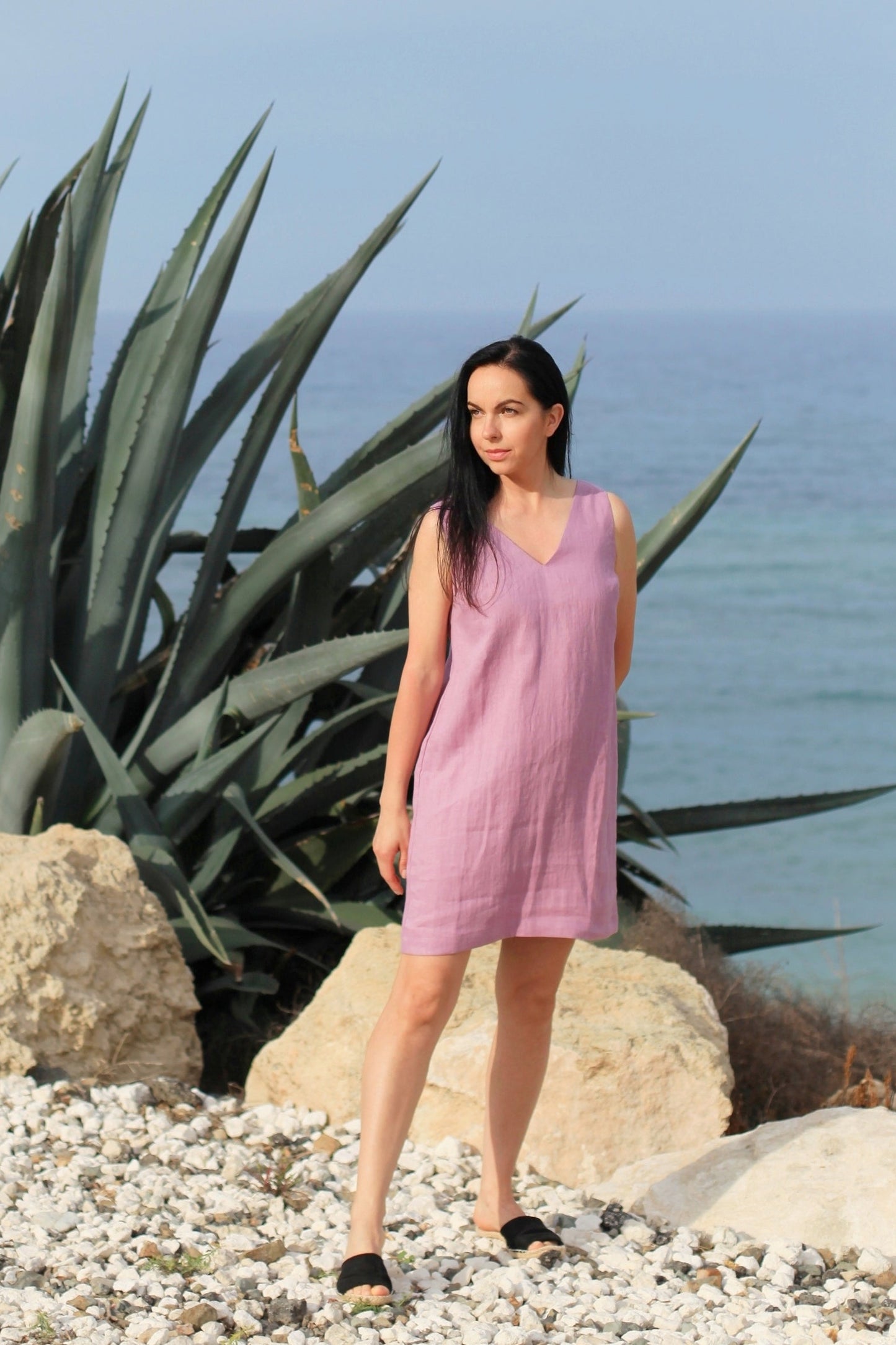 Woman in a pink dress standing on a pebbly beach with agave plants and ocean in the background