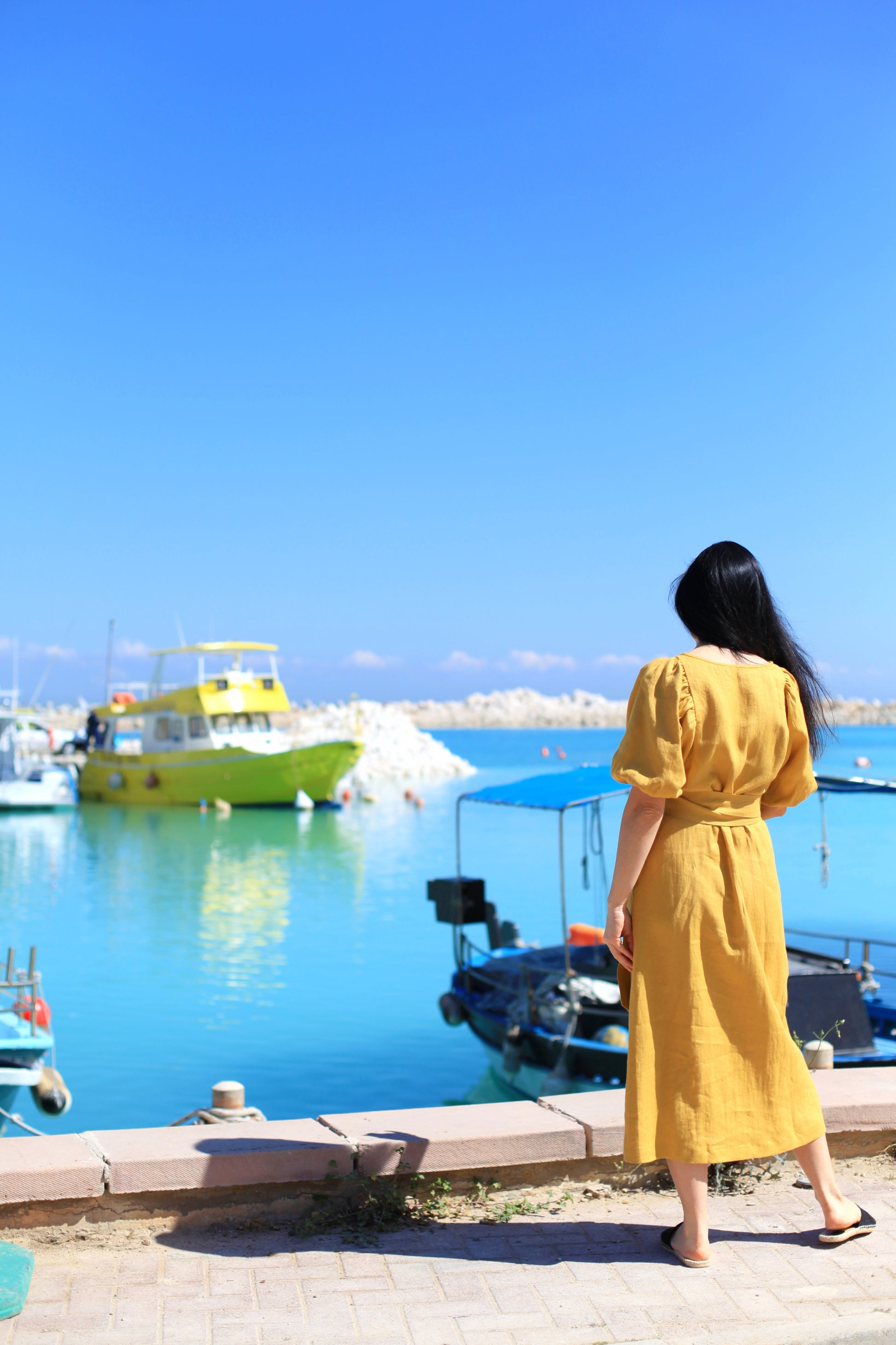 Woman in a yellow dress standing by a harbor with boats and clear blue sky.