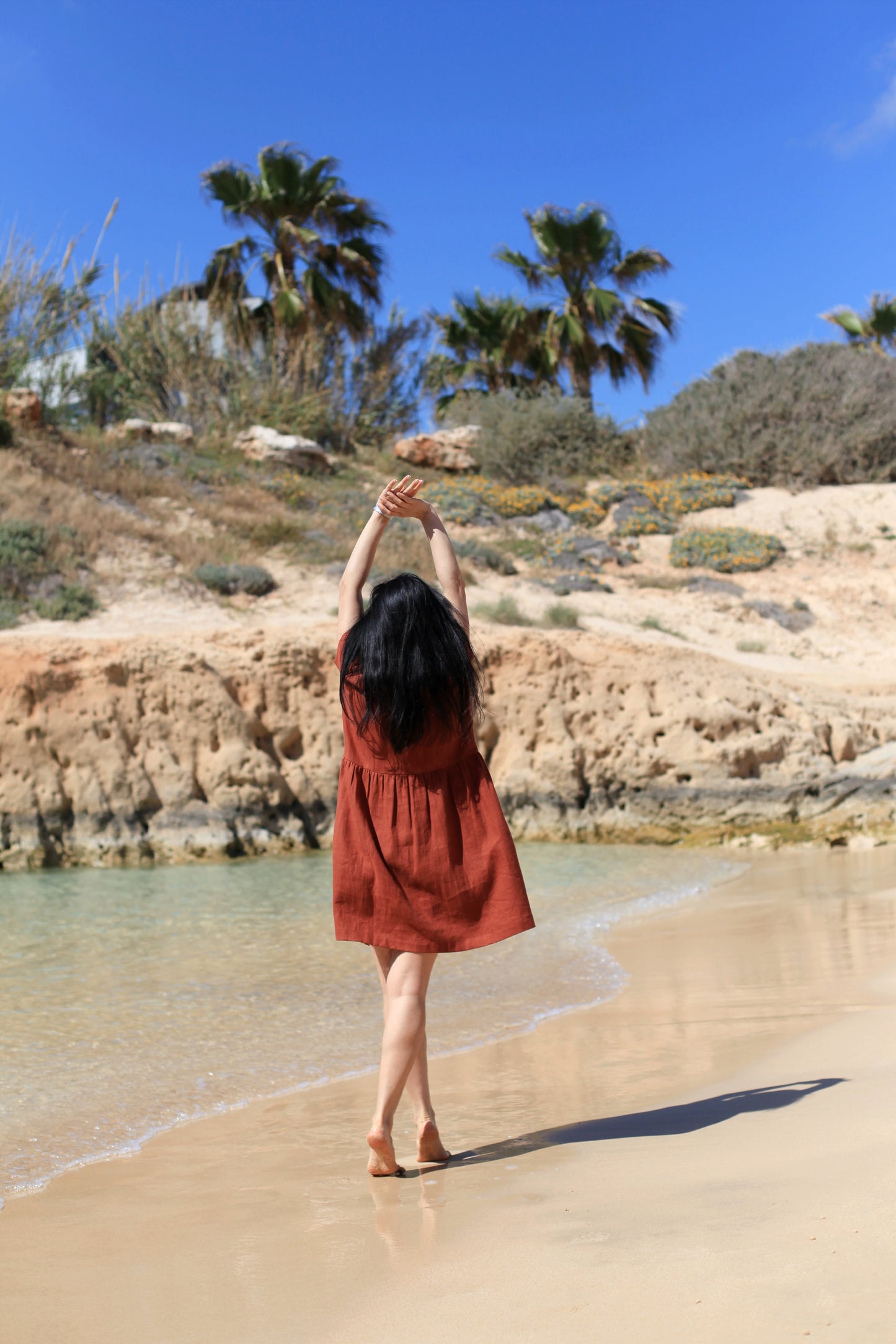 Woman in a red dress standing on a beach with palm trees and rocky cliffs in the background.