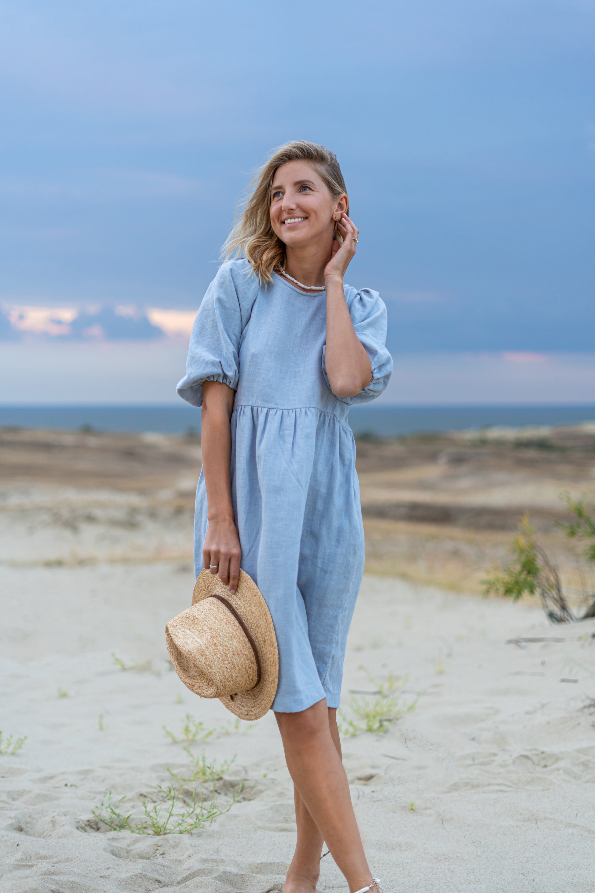 Light blue linen dress angled front pose with hat on beach, handmade linen dress slow fashion