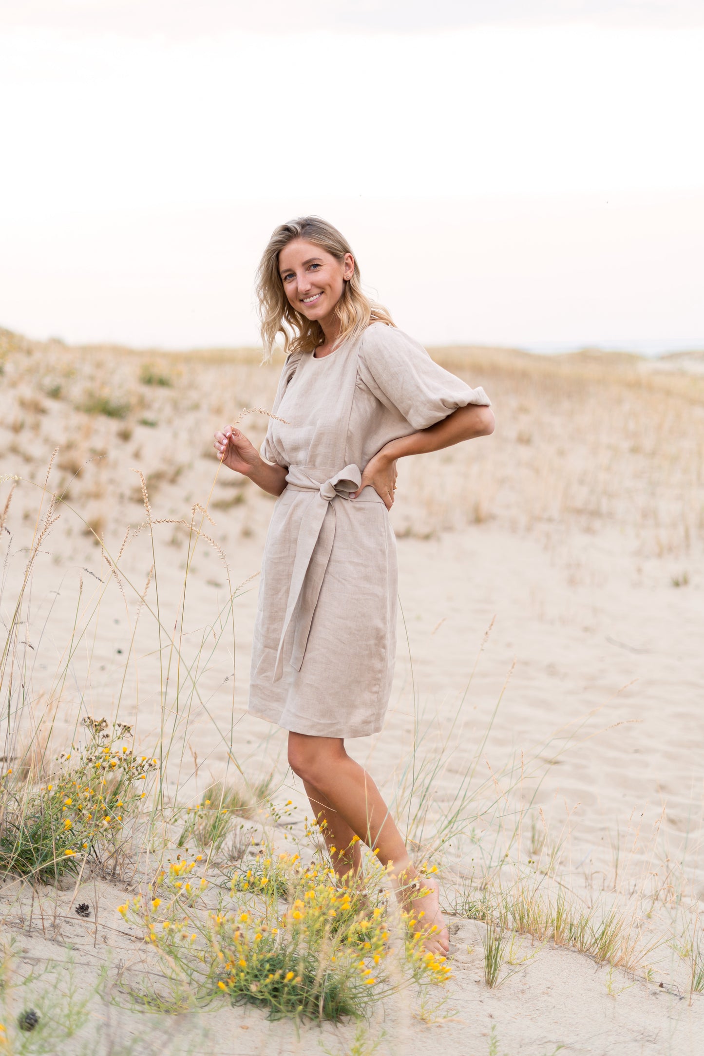 Woman in a beige dress standing in a sandy landscape with wildflowers.
