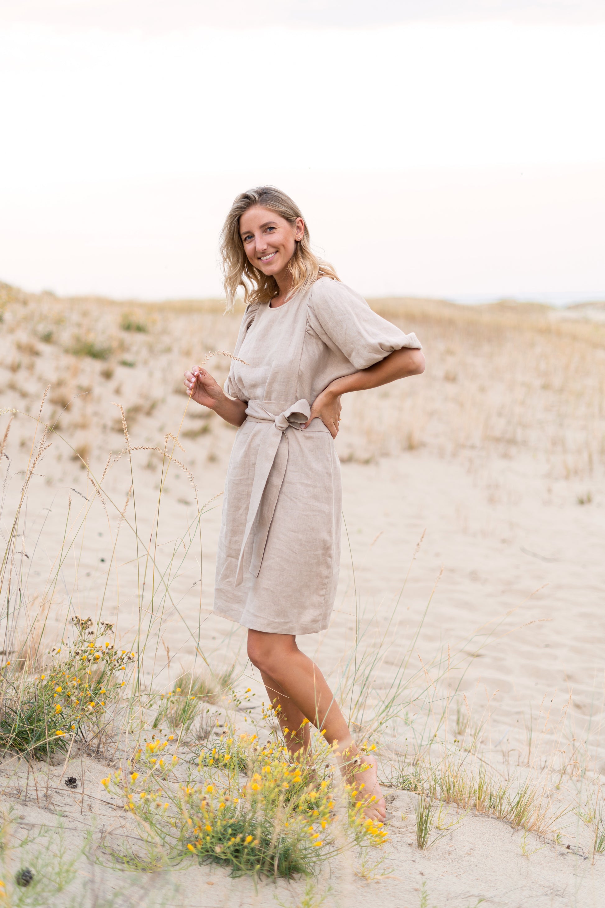 Woman in a beige dress standing in a sandy landscape with wildflowers.